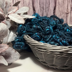 May include: A gray wicker basket filled with dark teal and blue yarn. The yarn has a textured, curly appearance. The basket is set against a blurred background of a wooden surface and decorative foliage.