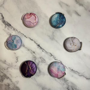 K&ouml;nnte beinhalten: Sieben runde Handyhalterungen mit Marmormustern und silberfarbenen Metallr&auml;ndern. Die Halterungen weisen verschiedene Farben auf, darunter Rosa, Blau, Lila und Wei&szlig;, mit goldenen Akzenten. Sie sind kreisf&ouml;rmig auf einer wei&szlig;-grauen Marmoroberfl&auml;che angeordnet.