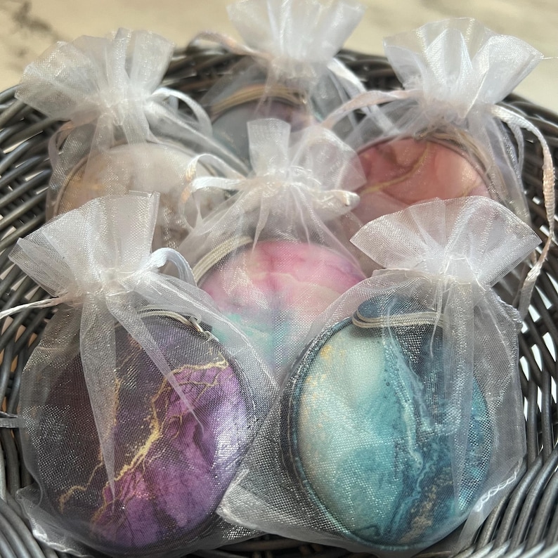 K&ouml;nnte beinhalten: Eine Sammlung runder, dekorativer Artikel mit abstrakten Designs in Lila, Blau, Rosa und Gold, jeweils in einem durchsichtigen wei&szlig;en Organzabeutel verpackt. Die Beutel sind mit einem Band gebunden und in einem grauen Weidenkorb angeordnet.