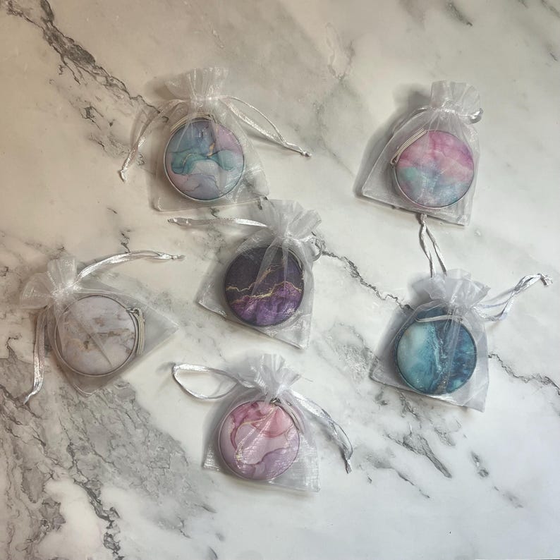 K&ouml;nnte beinhalten: Sieben runde Kompaktspiegel mit silbernen R&auml;ndern, jeweils in einem durchsichtigen wei&szlig;en Kordelzugbeutel verpackt. Die Spiegel zeigen farbenfrohe, abstrakte Designs in Blau-, Rosa-, Lila- und Wei&szlig;t&ouml;nen, die Marmor- oder Aquarellkunst &auml;hneln.