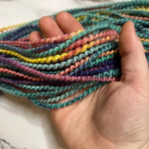 May include: A handful of colorful, textured yarn in shades of teal, pink, yellow, and purple. The yarn is tightly twisted, creating a spiral effect. The yarn is held in a hand, showcasing the vibrant colors and texture.