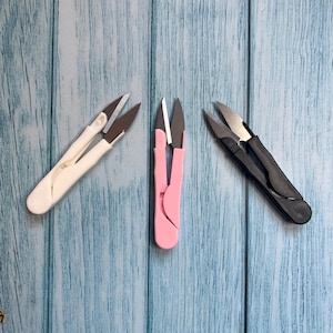 Könnte beinhalten: Drei Fadenscheren in Weiß, Rosa und Schwarz, angeordnet auf einer blauen Holzoberfläche. Jede Schere hat einen gebogenen Griff und scharfe, silberne Klingen. Diese kleinen Scheren sind für präzises Schneiden in Näh- und Bastelprojekten konzipiert.