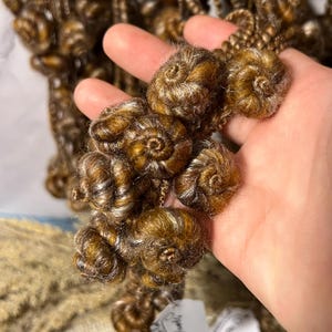 May include: Close-up of brown and blonde curly hair extensions. The hair is styled in small, tight curls, with a spiral shape. The hair has a mix of brown and blonde highlights, creating a natural look. The extensions are likely for braiding or adding volume.