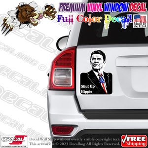 May include: A white car with a black and white sticker of a man with the text "Shut Up Hippie". The sticker is on the back window of the car.