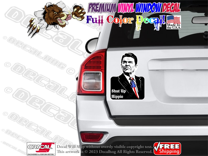May include: A white car with a black and white sticker of a man with the text "Shut Up Hippie". The sticker is on the back window of the car.