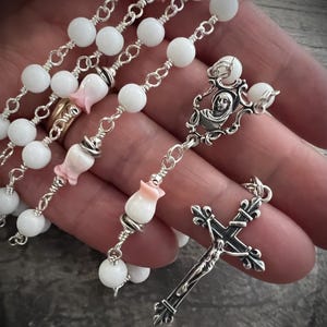Puede incluir: Un rosario con cuentas blancas y eslabones de cadena plateados. El rosario presenta cuentas en forma de flor rosa y blanca y un crucifijo plateado con una representación detallada de Jesús. El rosario se sostiene en una mano.