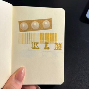 Könnte beinhalten: Notizbuchseite mit dekorativem Klebeband. Das Klebeband enthält drei perlenartige Kreise, vertikale Streifen und den Namen "KIM" in goldfarbenen Buchstaben. Die Seite wird von einer Hand mit hellrosa Nagellack gehalten.