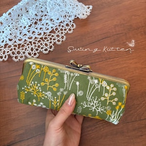 May include: A green clutch with embroidered yellow and white floral designs. The clutch has a gold-colored metal frame and clasp. The text "Swing Kitten" is written in cursive on the wooden surface.