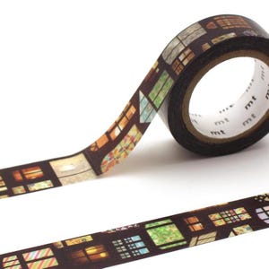 May include: A roll of decorative washi tape featuring a repeating pattern of colorful window illustrations. The tape is dark brown with a variety of window designs in shades of orange, green, and yellow. The tape is labeled with the brand name "mt".