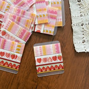 Könnte beinhalten: Dekorative Washi-Tape-Streifen mit rosa, gelben und roten Mustern. Die Designs umfassen Erdbeeren, Herzen und gewellte Kanten. Das Klebeband ist auf transparenten Plastikkarten angeordnet.
