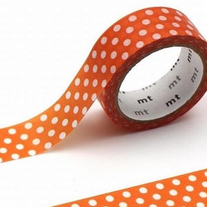 Könnte beinhalten: Orangefarbene Washi-Tape-Rolle mit weißen Polka-Dots. Das Klebeband ist teilweise abgerollt und zeigt den inneren Kern mit dem Aufdruck "mt". Dekoratives Bastelmaterial.