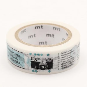 May include: A roll of washi tape with a vintage camera design. The tape is white with a light blue and black illustration of a camera and text that reads "OPERATING MANUAL CAMERA". The tape also has the text "mt" printed on it.