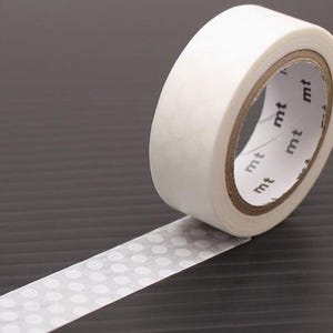 Könnte beinhalten: Eine Rolle weißes Washi-Tape mit einem sich wiederholenden Muster aus kleinen, weißen Punkten. Das Klebeband ist teilweise abgerollt und zeigt das gepunktete Design. Der Markenname "mt" ist auf dem Kern des Bandes aufgedruckt.