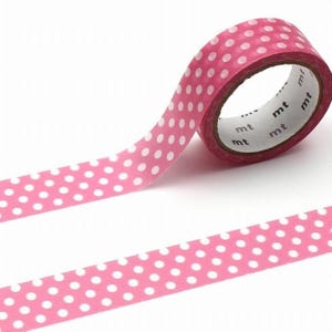 May include: A roll of pink washi tape with a white polka dot pattern. The tape is partially unrolled, revealing the pattern and the brand name "mt" printed on the core. The tape is designed for crafts and decorative purposes.