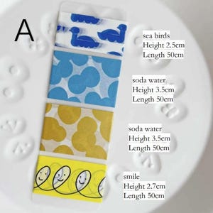 Könnte beinhalten: Vier dekorative Washi-Tape-Rollen mit verschiedenen Mustern und Texten. Die obere Rolle zeigt blaue Vögel, die zweite blaue Kreise, die dritte gelbe Kreise und die untere Rolle lächelnde Gesichter. Jede Rolle ist 50 cm lang.
