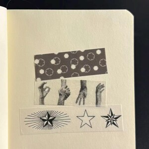 Könnte beinhalten: Eine Notizbuchseite mit dekorativem Washi-Tape. Das Tape zeigt ein Muster aus weißen Kreisen auf dunklem Hintergrund, Illustrationen von Händen und Sterndesigns. Das Notizbuch hat cremefarbenes Papier.
