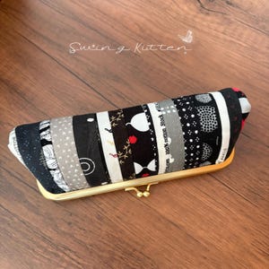 Peut inclure: Une pochette rectangulaire faite à la main avec un fermoir doré. Le tissu est un patchwork de bandes de coton à motifs noirs, blancs et gris. Le texte "Swing Kitten" est écrit en cursive en haut à gauche.