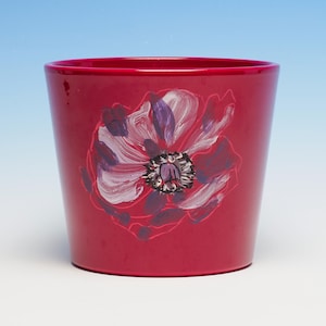 May include: A red ceramic plant pot with a hand-painted floral design. The flower has white, purple, and dark red petals with a dark centre. The pot has a glossy finish and a slightly tapered shape, suitable for indoor or outdoor use.
