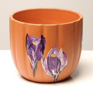 May include: A terracotta flower pot with a hand-painted design of purple crocuses. The pot has a rounded shape and a textured surface.