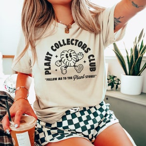 K&ouml;nnte beinhalten: Eine Frau tr&auml;gt ein beiges T-Shirt mit einer schwarzen Grafik, auf der "Plant Collectors Club" und eine Comic-Pflanzenfigur zu sehen sind. Der Text unter der Grafik lautet "Follow me to the Plant Store!"