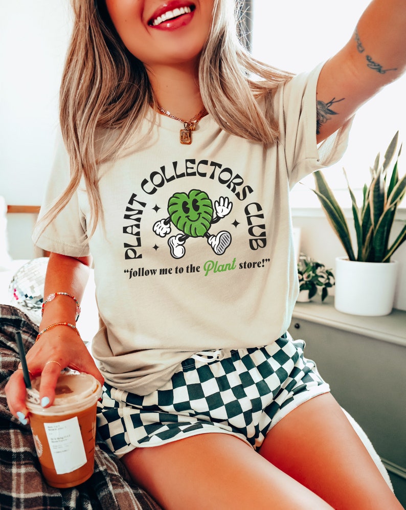May include: A woman wearing a light beige t-shirt with a green plant cartoon and the text "Plant Collectors Club" and the words "follow me to the Plant store!"  She is wearing black and white checkered shorts and holding a cup of coffee.
