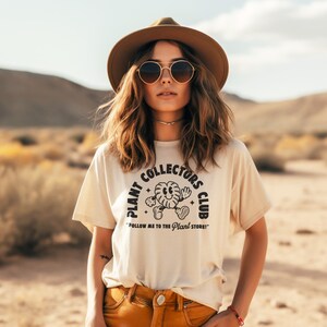 K&ouml;nnte beinhalten: Eine Frau tr&auml;gt ein beiges T-Shirt mit einer schwarzen Grafik, auf der "Plant Collectors Club" und eine Comic-Pflanzenfigur zu sehen sind. Der Text unter der Grafik lautet "Follow me to the Plant Store!"
