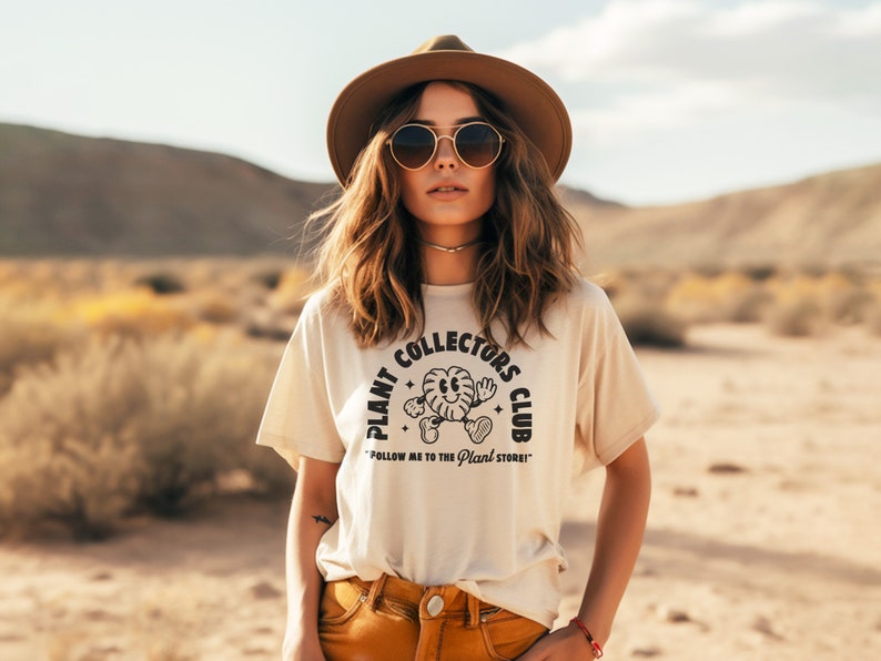 K&ouml;nnte beinhalten: Eine Frau tr&auml;gt ein beiges T-Shirt mit einer schwarzen Grafik, auf der "Plant Collectors Club" und eine Comic-Pflanzenfigur zu sehen sind. Der Text unter der Grafik lautet "Follow me to the Plant Store!"