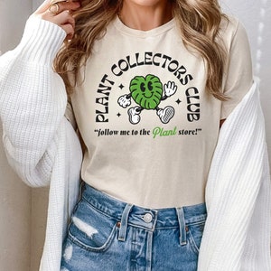 May include: A beige t-shirt with a green cartoon plant wearing a smile and holding a sign that says "Plant Collectors Club" and "follow me to the Plant store!"