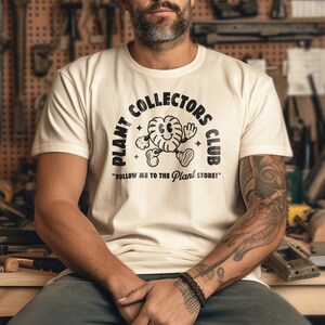 K&ouml;nnte beinhalten: Ein wei&szlig;es T-Shirt mit einem schwarzen Grafikdesign, das einen Comic-Charakter und den Text "Plant Collectors Club" und "Follow me to the Plant Store!" zeigt.