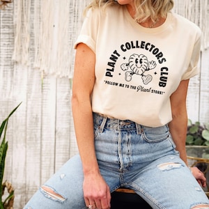 K&ouml;nnte beinhalten: Ein beiges T-Shirt mit einer schwarzen Grafik eines Cartoon-Pflanzencharakters und dem Text "Plant Collectors Club" und "Follow me to the Plant Store!"