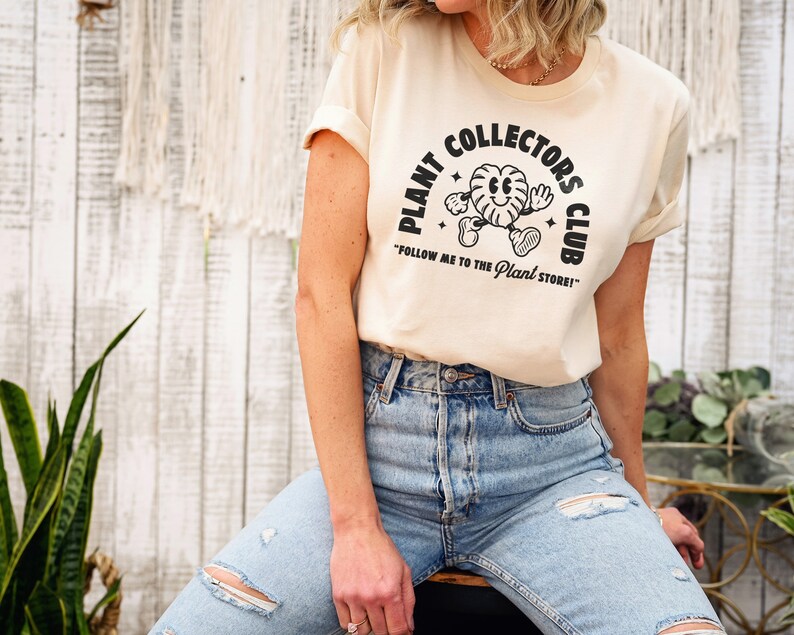 K&ouml;nnte beinhalten: Ein beiges T-Shirt mit einer schwarzen Grafik eines Cartoon-Pflanzencharakters und dem Text "Plant Collectors Club" und "Follow me to the Plant Store!"