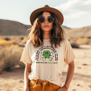 May include: A woman wearing a beige t-shirt with a green plant graphic and the text "Plant Collectors Club" and the words "Follow me to the Plant store!"  She is wearing a brown hat and sunglasses.