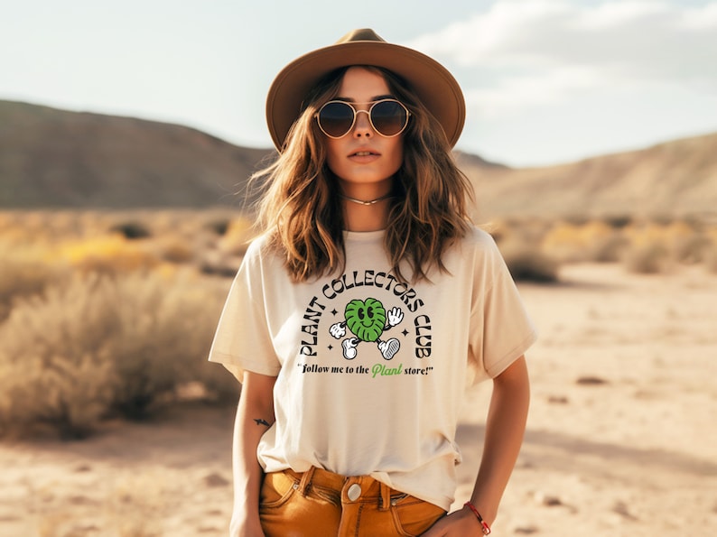 May include: A woman wearing a beige t-shirt with a green plant graphic and the text "Plant Collectors Club" and the words "Follow me to the Plant store!"  She is wearing a brown hat and sunglasses.