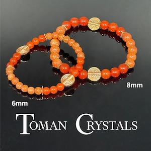 May include: Two orange beaded bracelets with gold-colored accents. One bracelet is labeled 6mm, the other 8mm. The bracelets are displayed on a reflective black surface. The text "Toman Crystals" is at the bottom.