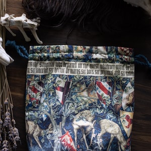 Pode incluir: Uma bolsa de cordão azul com um design de tapeçaria que apresenta dois veados brancos, uma árvore e uma faixa com o texto "ek the saugtral who departed on the quest whatroer might befal .sir lancelor sır hector or marys.sir bors, sir pririwal and Singalaga".