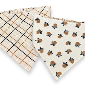 May include: Two dog bandanas, one with a brown and black plaid pattern and the other with a white background and brown acorn print.