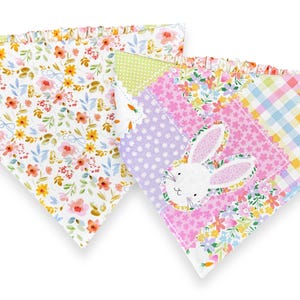 May include: Two colorful pet bandanas. One features a floral pattern with yellow and orange flowers. The other has a patchwork design with a bunny face, polka dots, and floral prints. Both have ruffled edges.