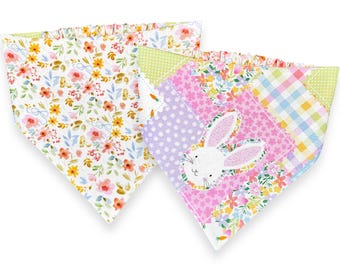 May include: Two colourful pet bandanas. One features a floral pattern with yellow and orange flowers. The other has a patchwork design with a bunny face, polka dots, and floral prints. Both have ruffled edges.