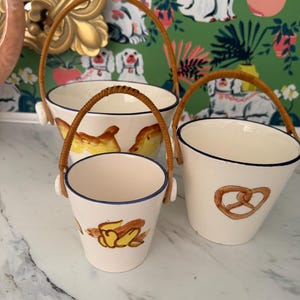 May include: Three off-white ceramic buckets with brown woven handles. Each bucket has a blue rim and a different food design: bread, a pretzel, and a banana. The buckets are on a white marble surface.