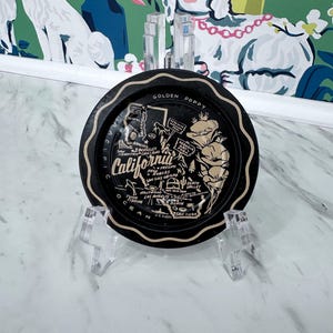 May include: Black and gold metal souvenir plate featuring a map of California with the text "Golden Poppy" and "Pacific Ocean".