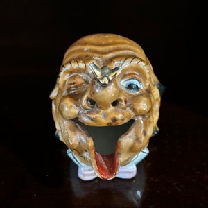 May include: A ceramic ashtray shaped like a man's face with a winking eye and a butterfly on his forehead. The ashtray has a red tongue and a white collar.