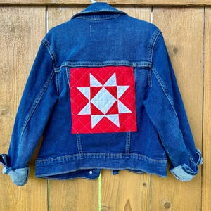 May include: A blue denim jacket with a red and white star quilt block design on the back. The jacket is hanging on a white hanger against a wooden fence background. The sleeves are cuffed.
