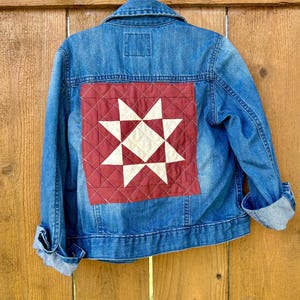 May include: A blue denim jacket with a red and white star quilt patch on the back. The jacket is hanging on a white hanger against a wooden fence background. The sleeves are cuffed.