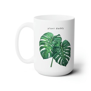 Pode incluir: Caneca de cerâmica branca com um design de folha de monstera verde e o texto "plant daddy" impresso na frente.
