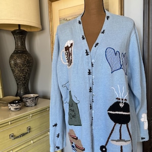 May include: Light blue cardigan sweater with embroidered barbecue-themed designs. Features a beer mug, oven mitts, an apron with "Kiss the Chef" text, a grill, and steaks. The sweater has a button-up front and small black accents.