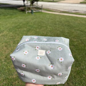 May include: A sage green fabric pouch featuring a repeating pattern of white daisies with yellow centers. The pouch has a white zipper closure along the top and a small rectangular beige label centered on the front with illegible text.