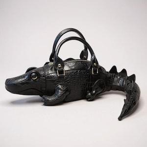 May include: A black alligator-shaped handbag with a textured, scaly design. It has two top handles, a zip closure, and a tail. The alligator's eye is yellow.