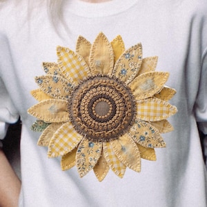 Puede incluir: Sudadera blanca con un gran diseño de girasol aplicado. El girasol tiene pétalos amarillos de cuadros y estampados florales, un centro marrón y está cosido en la parte delantera. La sudadera tiene mangas largas y cuello redondo.