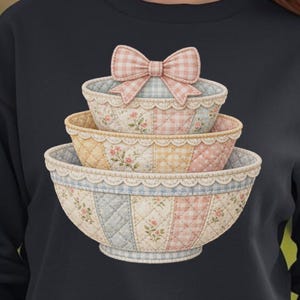 May include: Black sweatshirt featuring a stack of three decorative bowls. The bowls have a quilted pattern in pastel colors: blue, pink, and yellow. Each bowl is adorned with floral designs and a lace trim. A pink gingham bow sits atop the stack.