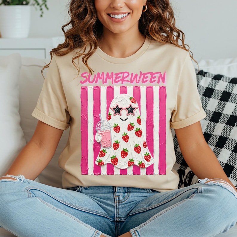 May include: Beige t-shirt with the word "SUMMERWEEN" in pink. The shirt features a ghost design wearing sunglasses and covered in strawberries, holding a pink drink. The background has pink and white stripes.